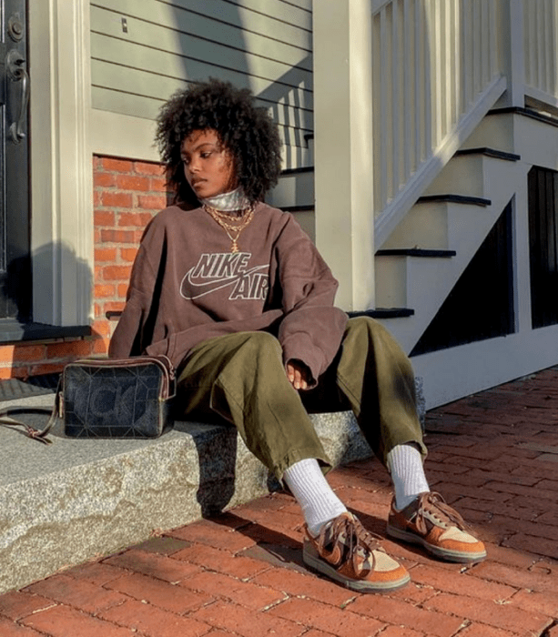 chica morena de cabello chino usando una sudadera oversized café, pantalón de pana verde militar, tenis naranjas, bolso de piel negro