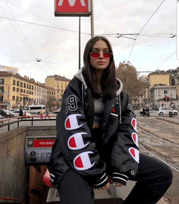 chica de cabello castaño usando lentes de sol rojos, top gris, chamarra negra oversized con logos grandes, mom jeans negros