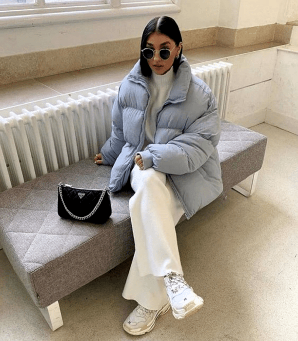 chica de cabello corto usando lentes de sol, suéter blanco, chamarra puffer jacket azul celeste, pantalones blancos holgados, tenis blancos y bolso negro