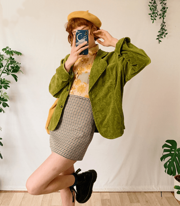 chica pelirroja con un beret amarillo, camisa amarilla con flores, abrigo verde de terciopelo, minifalda beige de cuadros y botas negras de suela gruesa