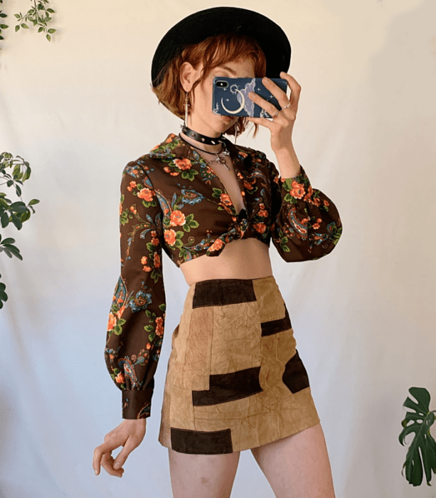 chica pelirroja usando un sombrero negro de ala ancha, crop top negro de flores con manga larga, minifalda beige con cuadros cafés