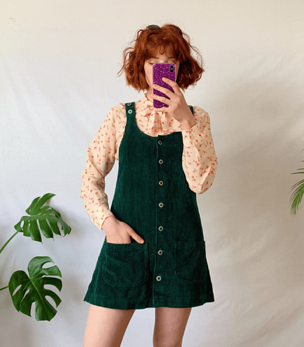 chica pelirroja usando un vestido verde de pana estilo overol, blusa beige de manga larga con estampado de flores rosas