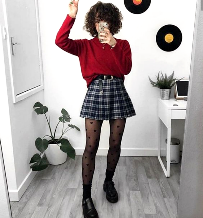 chica de cabello chino usando un suéter rojo de manga larga, minifalda de tablones escocesa con cuadros azules y blancos, medias negras con corazones, cinturón negro y mocasines negros de suela gruesa