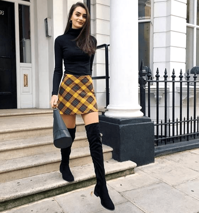 chica de cabello castaño largo usando un top negro de cuello alto y manga larga, minifalda de cuadros escocesa color amarillo con azul, botas largas negras de tacón bajo y bolso de mano de cuero