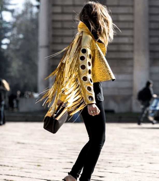 chica de cabello castaño usando una chamarra dorada metálica de manga larga con perforaciones en las mangas, skinny jeans negros, top negro y bolso de mano negro