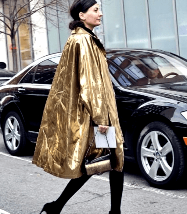 chica de cabello castaño usando un abrigo largo dorado metálico, bolso de mano negro con dorado metálico, medias negras gruesas y zapatos de tacón negros