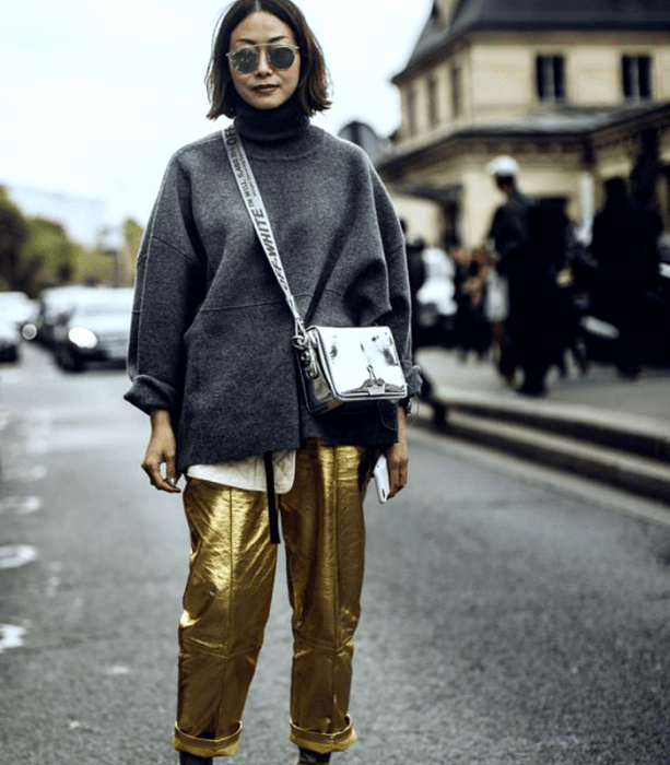 chica de cabello castaño usando lentes de sol, sudadera gris de cuello alto, bolso de mano metálico plateado, pantalones dorados metálicos