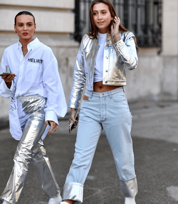 chicas de cabello castaño, una está usando una camiseta blanca de vestir con estampado, pantalones metálicos plateados y botas blancas, la otra usa una camiseta blanca crop top, chaqueta metálica plateada, jeans a la cadera con vistas metálicas y botas blancas