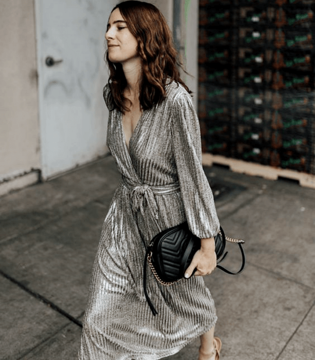 chica de cabello castaño usando un vestido largo metálico plateado, bolso de mano negro y sandalias de tacón beige