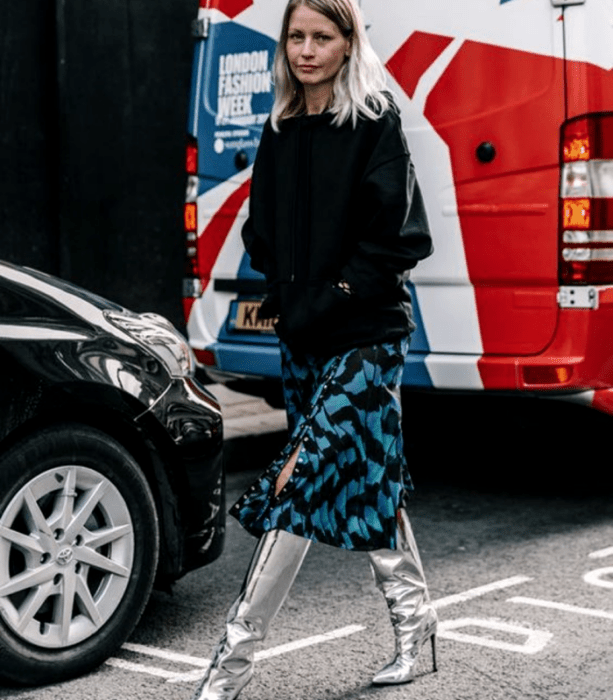 chica rubia usando un top negro de manga larga, falda midi azul con lineas negras, botas largas metálicas plateadas