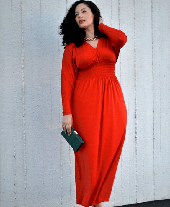 chica curvy de cabello chino usando un vestido rojo largo de cuello en V, manga larga y bolso de mano negro