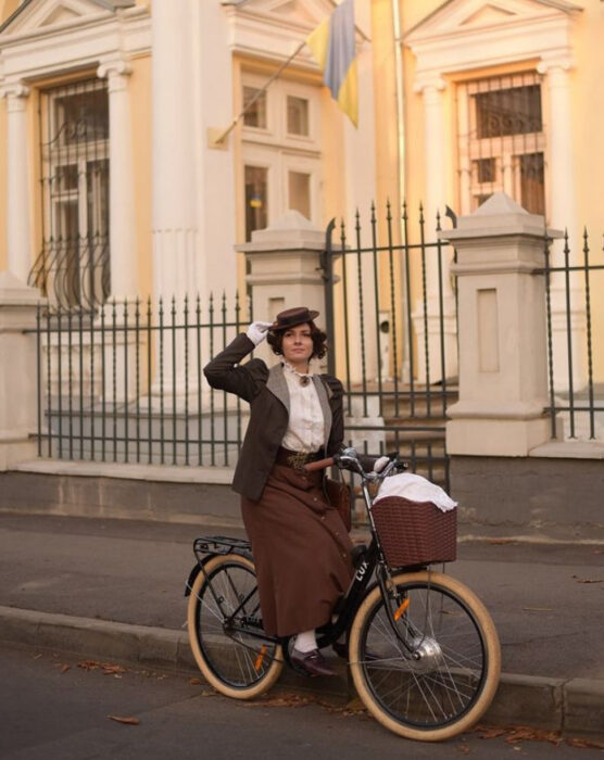 Mila Povoroznyuk usando atuendo de época, de falda larga color café, blusa blanca y saco gris, con cabello recogido y sombrero, montando una bicicleta