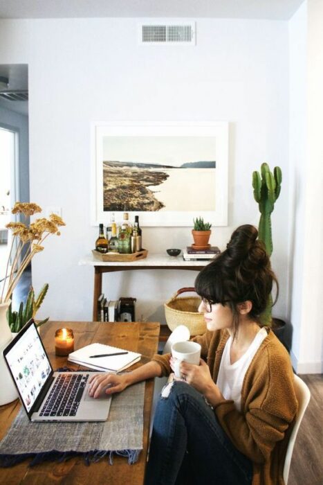 Chica trabajando en la computadora, su estudio tiene varias plantas en el lugar, como un cactus o una platita pequeña cerca de la computadora