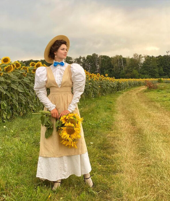 Mila Povoroznyuk usando vestido de época color blanco con ensamble color caki y cargando unos girasoles 