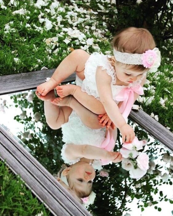 Bebé usando una diadema, con vestido blanco y sentada sobre un espejo viendo su reflejo