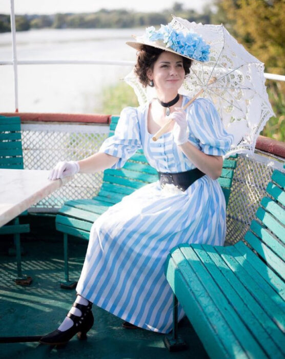 Mila Povoroznyuk usando atuendo de época, de vestido de rayas azul cielo con blanco, cinto negro, guantes blancos, sombrero y sombrilla