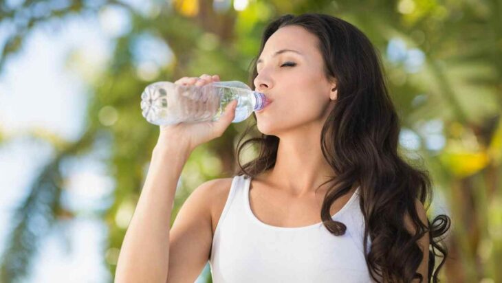 Chica morena, delgada y de cabello largo ondulado con blusa blanca tomando agua de una botella pequeña de plástico 