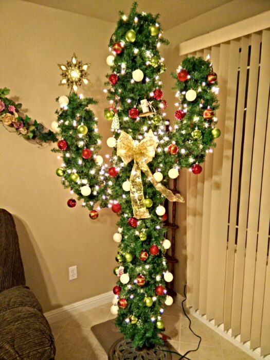 Árbol de navidad en forma de cactus colocado en una sala con paredes y persianas blancas decorado con luces amarillas, esferas rojas y un moño grande dorado