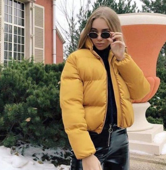 Chica usando chamarra de color mostaza, con jeans y blusa negra y lentes de sol