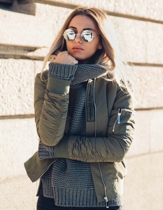 Chica usando caqueta bomber color verde militar con suéter gris debajo y lentes de sol