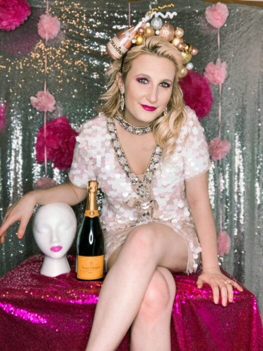 Blonde Frau, die am Tisch mit rosa Tischdecke sitzt und weißes Kleid mit V-Ausschnitt und Geburtstagskrone mit Flasche Champagner und Blasen trägt 