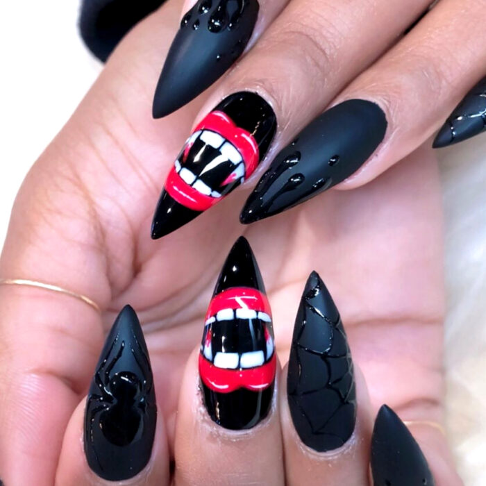 Manos con uñas largas stiletto pintadas con esmalte negro mate, con diseño de arañas, telaralas y labios rojos con colmillos de vampiro