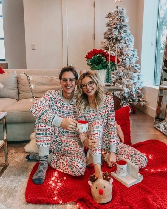 Pareja sentada en el suelo, usando la misma pijama y con la casa decorada de Navidad