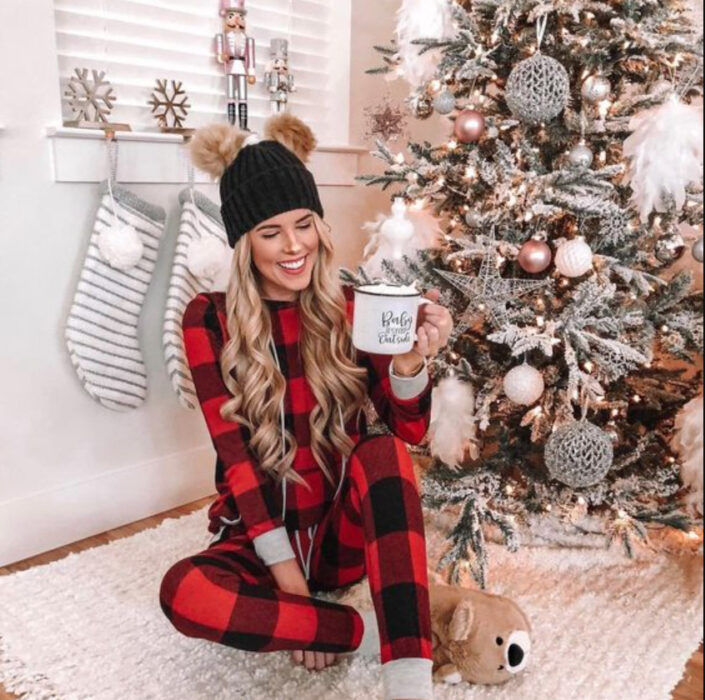 Chica usando pijama de navidad, sentada sobre el suelo, delante del árbol de navidad bebiendo una taza de chocolate caliente