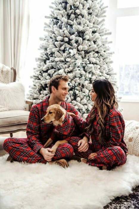 Pareja sentados sobre el suelo, usando la misma pijama posando delante de su árbol de navidad y acompañados de su perro