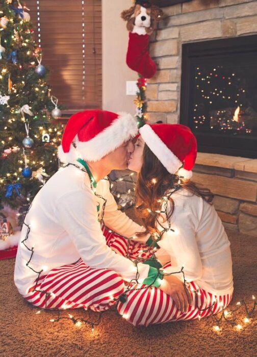 Pareja sentada de frente con el árbol de navidad detrás de ellos, besándose rodeados con luces navideñas