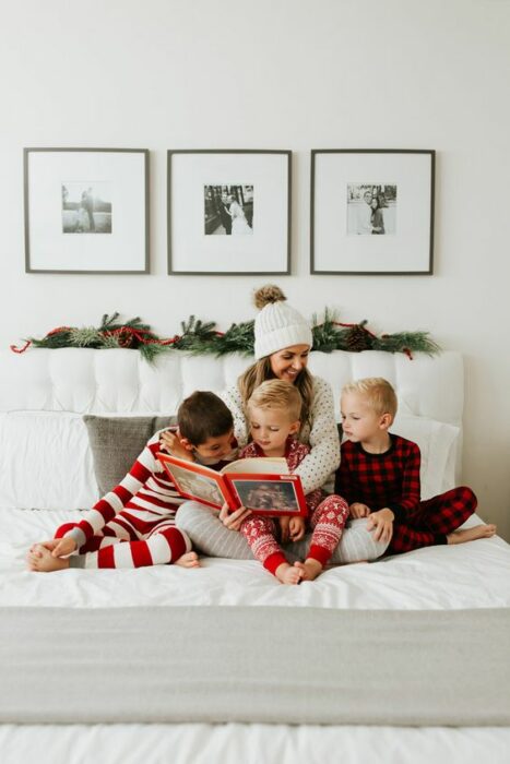 Chica con tres niños, todos sentados sobre la cama usando pijamas de navidad, mientras leen un cuento