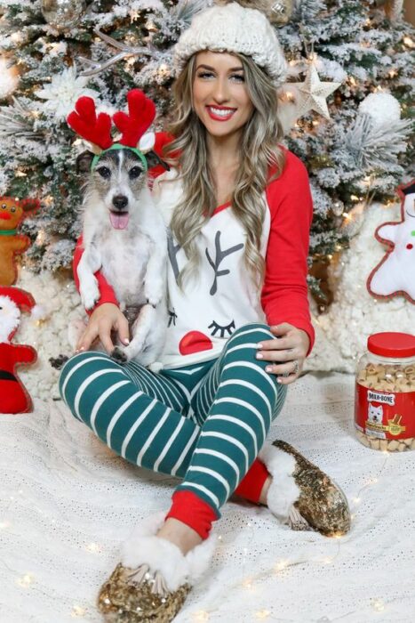 Chica usando pijama de navidad, sentada delante del árbol navideño, acompañada de su perro que usa una diadema de reno