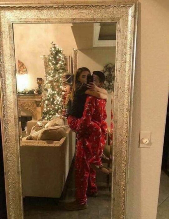 Das Paar macht ein Selfie vor dem Spiegel, während er einen roten Pyjama trägt, während er ein langhaariges blondes Mädchen hält