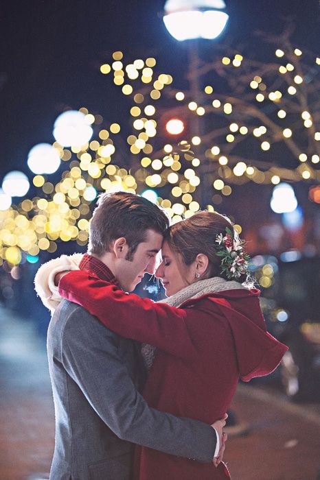 Hombre con abrigo gris abraza por la cintura a chica con cabello recogido en chongo y saco rojo con luces navideñas de fondo