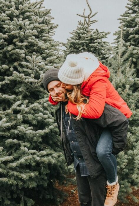 Hombre con gorro gris y chamarra negra carga en su espalda a chica rubia con gorro blanco y chamarra roja con pinos navideños de fondo