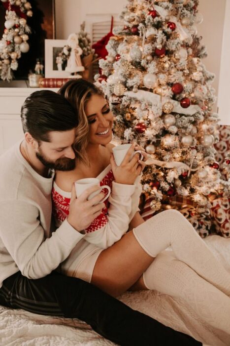 Mujer rubia de cabello largo sentada encima de su novia con suéter blanco ambos con tazas blancas de café en la mano sentados junto al árbol navideño