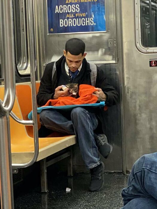 Chico sentado en el metro con saco negro, camisa blanca, corbata negra y jeans con una mochila gris en la espalda, alimenta a gatito café con rayas negras mientras lo arropa con una manta naranja y le da de comer con un biberón 