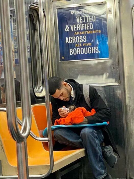 Chico sentado en el metro con saco negro, camisa blanca, corbata negra y jeans con una mochila gris en la espalda, alimenta a gatito café con rayas negras mientras lo arropa con una manta naranja y le da de comer con un biberón 