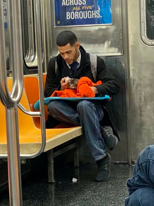 Chico sentado en el metro con saco negro, camisa blanca, corbata negra y jeans con una mochila gris en la espalda, alimenta a gatito café con rayas negras mientras lo arropa con una manta naranja y le da de comer con un biberón 