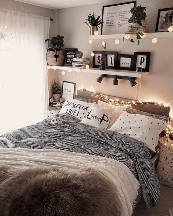 Habitación con paredes blancas con repisas en la pared con cuadros y libros encima y una cama en medio con cobija gris y cojines blancos con puntos negros y serie de luces amarillas para dar luz