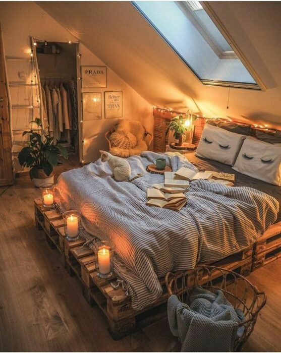 Habitación acogedora con tragaluz abajo de la cama que tiene base de pallets y cobijas grises con cojines blancos y paredes blancas también