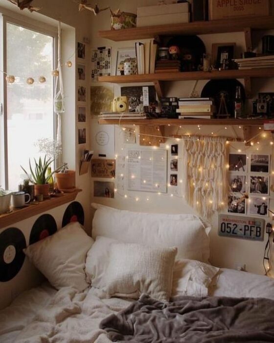 Habitación acogedora con cama en una esquina pegada la ventana con cobijas y cojines blancos y la pared blanca con repisas de madera llenas de discos y libros y series de luces amarillas colgando