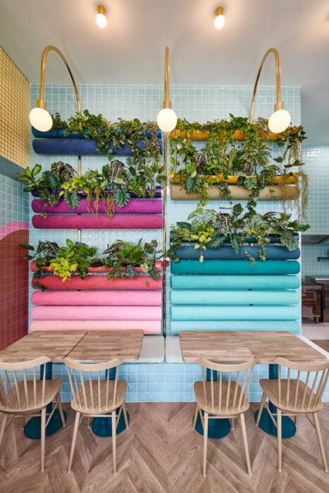Pared de restaurante color azul con macetas rectangulares de colores con mesas y sillas redonda de madera