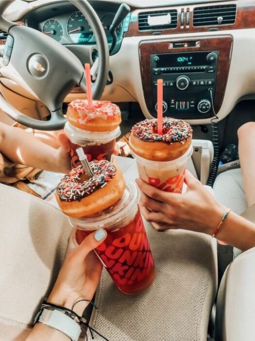 Manos sosteniendo bebidas con popotes y arriba unas donas de chocolate con chispas de colores