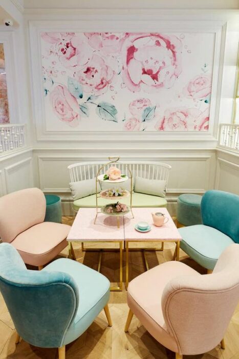 Restaurante con paredes blancas con tapiz de flores rosas con ramas verdes y en el centro de la habitación cinco sillones de colores blancos, azules y rosas con una mesa rectangular rosa en medio