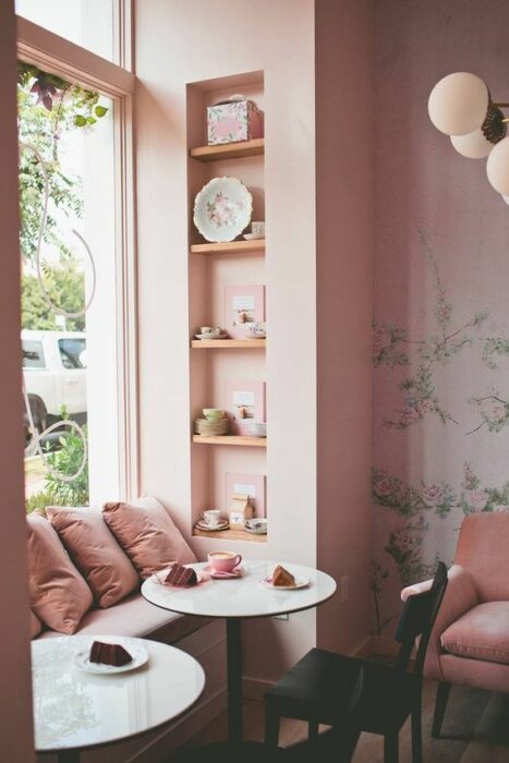 Cafetería color rosa con ventanal grande que da directo a la calle con asiento decorado con tres cojines rosa y una mesa pequeña redonda en medio 