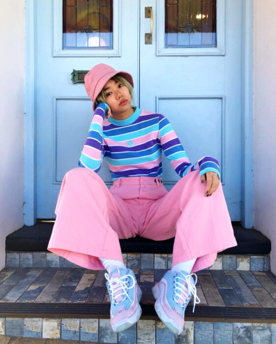 Looks coloridos aesthetic; mujer coreana de cabello corto y rubio sentada en las escaleras frente a una puerta azul, con ropa de colores, gorro pescador rosa, suéter rayado morado, azul fuerte y claro, pantalón acampanado y tenis chunky en tonos pastel