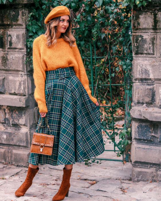 Looks coloridos aesthetic; mujer sonriendo con atuendo de colores, boina francesa y suéter oversized tejido anaranjados, falda larga con cuadros escoceses verde, bolsa de mano pequeña café, botines de gamuza y tacón, cabello ombré castaño en las raíces degradado a rubio en las puntas 