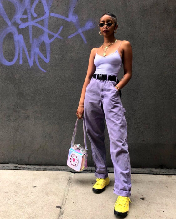 Looks coloridos aesthetic; mujer afroamericana de piel morena, cabello corto, lentes de sol pequeños, blusa de tirantes lila, mom jeans, pantalones de mezclilla morados, tenis amarillos con negro, cinto, bolsa de mano en forma de teléfono antiguo de colores holográficos