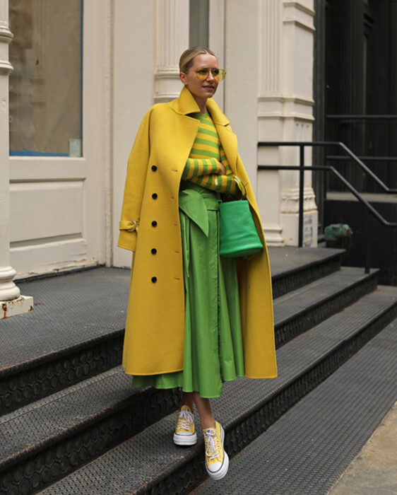 Looks coloridos aesthetic; mujer tomándose fotos en escaleras y frente a puerta blanca, cabello rubio recogido en una coleta baja, lentes de sol amarillos, saco largo, gabardina, blusa de manga larga con rayas amarillas y verdes, falda moderna, tenis converse, bolsa de mano